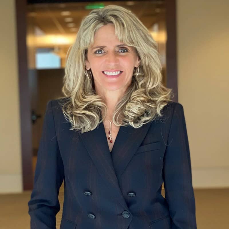 A smiling woman (Dina Barmasse-Gray) with long, wavy blonde hair, wearing a dark navy blazer over a black shirt. She has hoop earrings and a necklace with a red pendant. She is standing indoors with a modern office background.