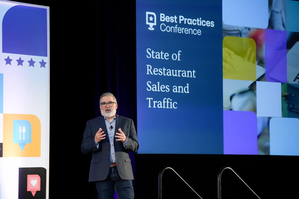 A man stands on a stage giving a presentation at the "Best Practices Conference." A large screen behind him displays the text "State of Restaurant Sales and Traffic" alongside various images collaged together.