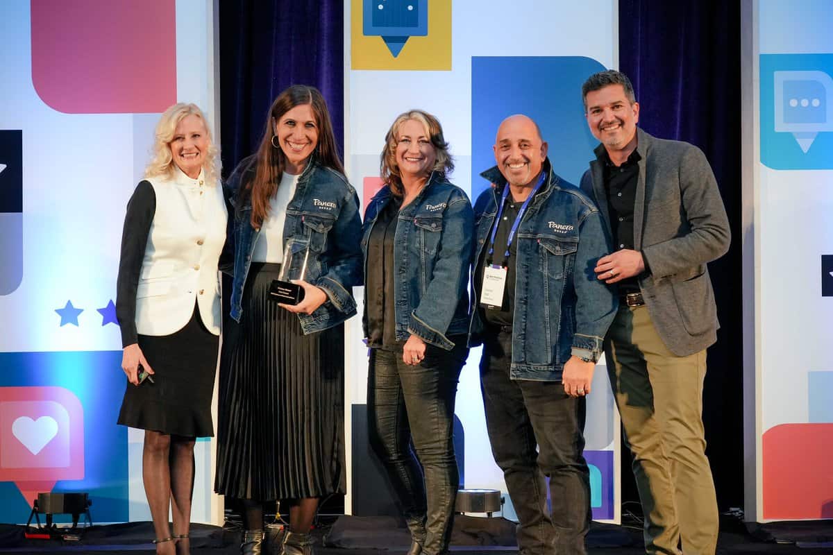 A group of five people smiling and posing on stage. Three individuals in the middle are wearing blue denim jackets, one of whom is holding a trophy. They are flanked by two others dressed in professional attire. The background features colorful, geometric designs.