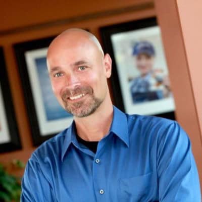 A smiling bald man (Matt Klug) with a short beard and mustache is wearing a blue button-up shirt and standing in front of a blurred background with framed photos. His arms are casually resting in front of him.