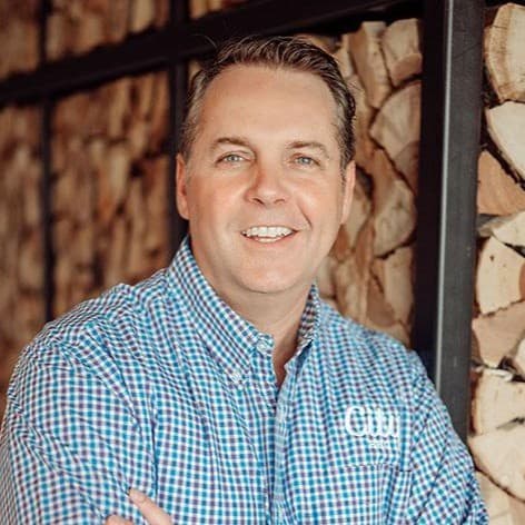 A man (Ronnie Berry) in a checkered shirt smiles while standing in front of a wall stacked with firewood logs. The logo on his shirt reads "CW". He has short, dark hair and is looking directly at the camera, exuding a friendly and approachable demeanor.