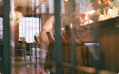 A softly lit café with large windows, wooden chairs, and people sitting at tables and at the bar, some blurred in motion, creating a warm and lively atmosphere.