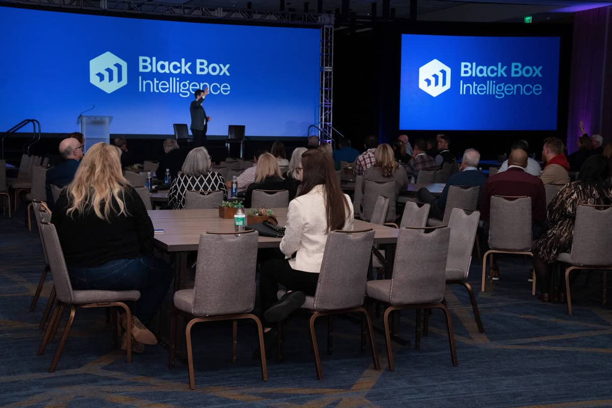 A speaker is presenting on stage at a conference with a large screen behind them displaying the "Black Box Intelligence" logo. Attendees sit at round tables, listening attentively. The room is dimly lit with a focus on the screen and the presenter.