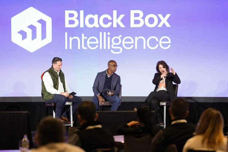 Three individuals are seated on stage engaged in a panel discussion in front of an audience. The backdrop displays the "Black Box Intelligence" logo. One person is speaking animatedly, another looks attentive, and the third holds a tablet.