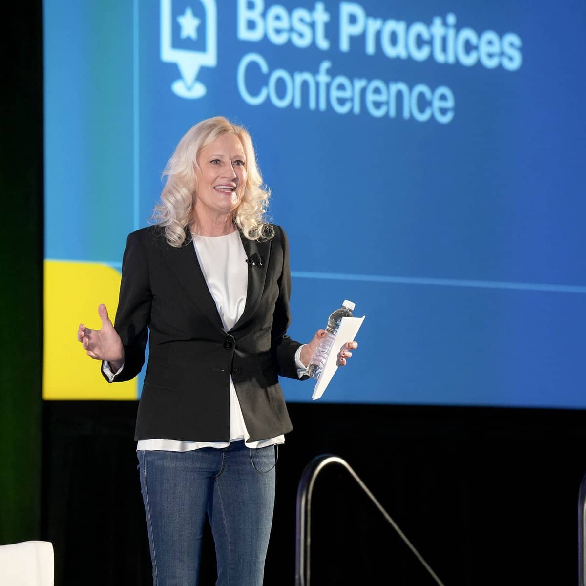 A speaker with long blonde hair (Mary Hamill), dressed in a black blazer and jeans, stands on stage at the Best Practices Conference. They hold a microphone and gestures while smiling in front of a blue and yellow conference backdrop.