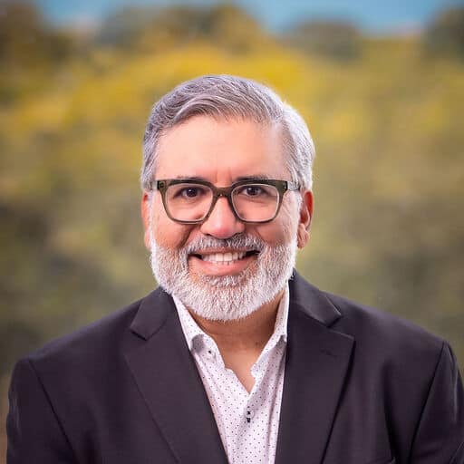 A man with gray hair and a beard, wearing glasses, a dark suit jacket, and a white shirt, smiles at the camera. The blurred green and yellow background suggests he's ready to discuss the State of Restaurant Industry Q2.