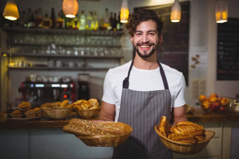 A smiling man wearing an apron stands in a bakery, holding baskets filled with various breads and pastries. The background shows shelves with glassware and a menu board. Soft lighting from hanging lamps creates a warm atmosphere.