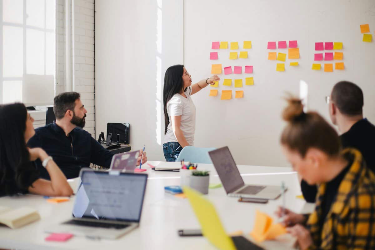 A group of people in a bright office participate in a brainstorming session. A woman stands at a whiteboard with colorful sticky notes. Others sit at a table with laptops, attentively listening and taking notes.