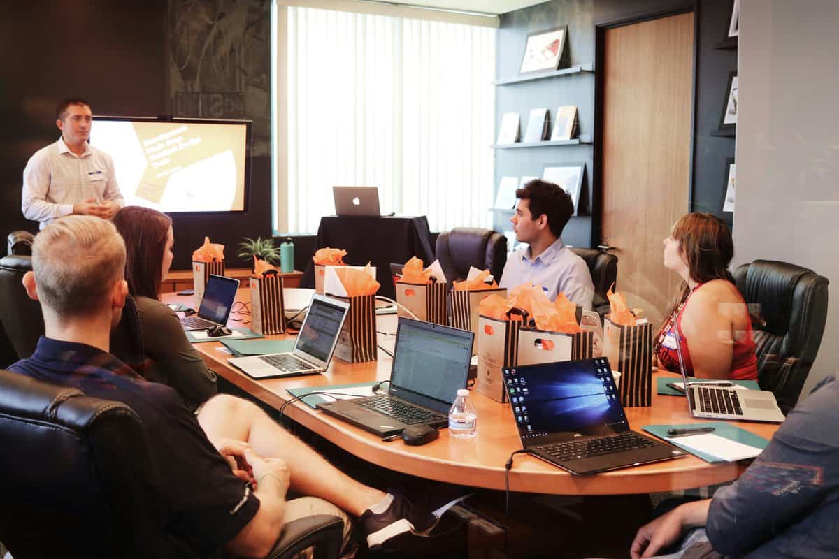 A man presents to a diverse group sitting around a conference table with laptops and gift bags. A screen displays his presentation. The room has bookshelves and large windows in the background.