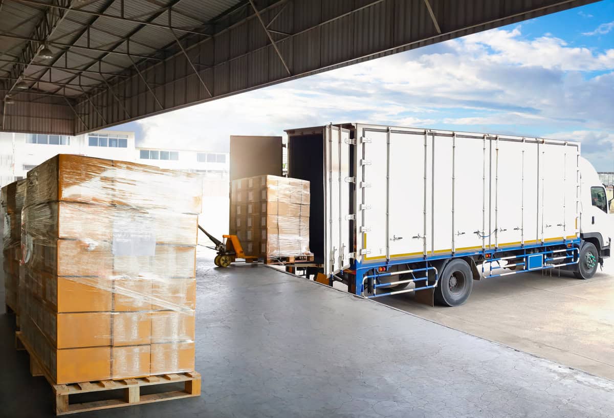 A white delivery truck is being loaded with large cardboard boxes on wooden pallets, using a pallet jack, at a warehouse dock. The area is partially covered, with an open view of the sky and nearby buildings.