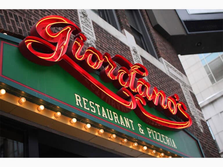 A restaurant sign for "Giordano's" with bright red, cursive lettering above green and yellow text that reads "Restaurant & Pizzeria." The sign is adorned with light bulbs and mounted on a brick building.