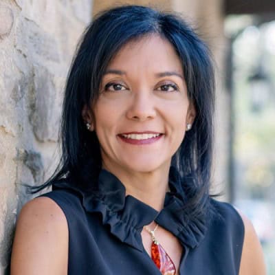 Image of Arlene Petokas Smiling woman with shoulder-length black hair, wearing a sleeveless black top with a ruffled collar. She's standing outdoors against a stone wall, wearing small earrings and a necklace with a reddish pendant. Background is softly blurred.