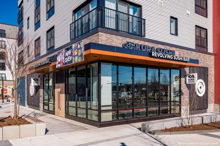 Street view of Kura Sushi, a revolving sushi bar with a modern exterior featuring large glass windows and entrance. The building is surrounded by a clean sidewalk and minimal landscaping, set in a contemporary urban area.