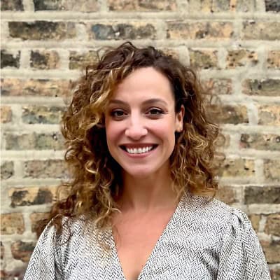 A person with curly hair is smiling while standing in front of a brick wall. She is wearing a light patterned top