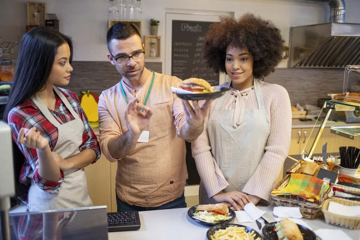 Three people stand behind a counter in a cafe. A man in glasses points at a burger on a plate he's holding, while two women in aprons watch. Plates with fries and sandwiches are on the counter.
