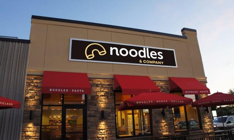 Exterior of a Noodles & Company restaurant with a modern facade. Features red awnings labeled "Noodles - Pasta" over windows and entrances. Store sign is prominently displayed above, showing a logo with stylized noodles.