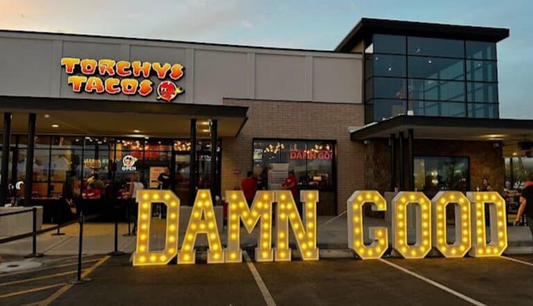 Exterior of Torchy's Tacos restaurant with a lighted sign saying "DAMN GOOD" in front. The building is modern with large windows, and the entrance is decorated with lights and outdoor seating. The sky is clear at dusk.