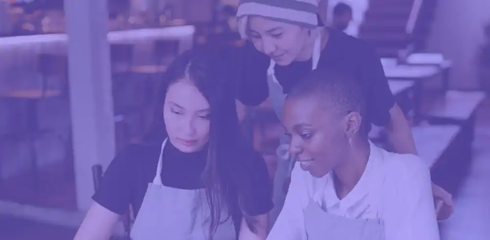 Three people wearing aprons are seated at a table in a restaurant, collaborating and looking at something in front of them. One person stands behind, leaning in to join the conversation. The image has a purple overlay.