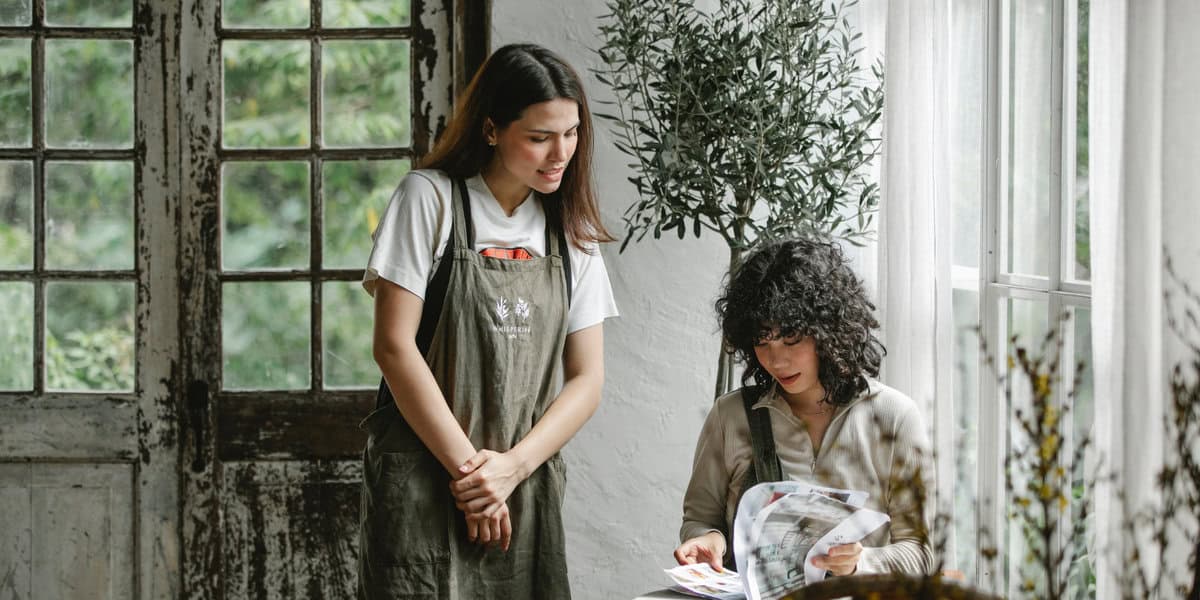 Two women are in a bright room; one stands wearing an apron and smiles, perhaps pondering how to deal with disgruntled customers, while the other sits by a window reading a magazine. Natural light streams in, and plants decorate the space.