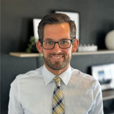 A man with short brown hair, a beard, and glasses is smiling. He is wearing a light-colored shirt and plaid tie. The dark wall behind him displays shelves with framed pictures, a decorative item, and hints of financial intelligence nextgen demo materials.