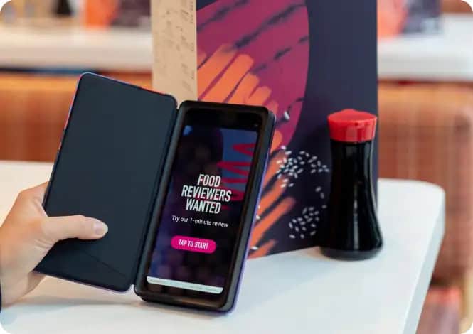 A person holds a smartphone displaying "Food Reviewers Wanted" next to a closed menu and a soy sauce dispenser, inviting participation in Restaurant Survey Collection.