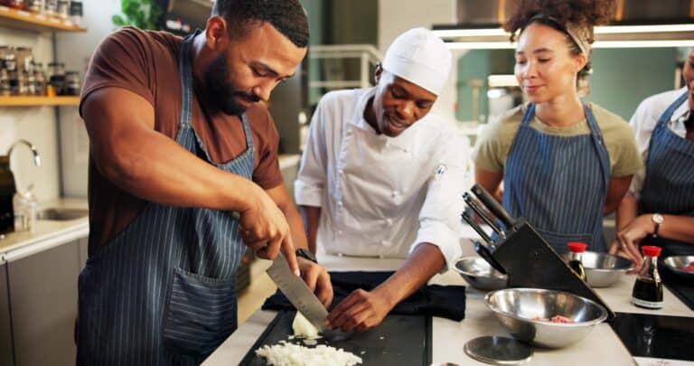 Three people in a kitchen, two in aprons and one in a chef’s uniform, watch as onions are chopped on a cutting board. The chef gives guidance, reflecting the teamwork often seen in diverse restaurant employee demographics.