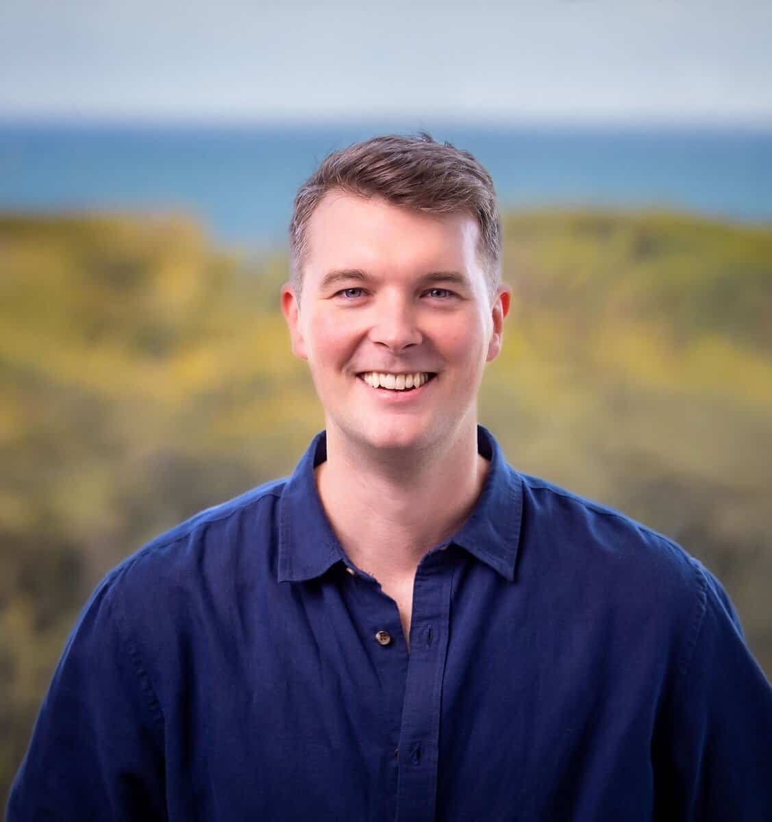 A man with short brown hair, wearing a blue button-up shirt, stands outdoors smiling. The background shows a blurred landscape with greenery and water in the distance.