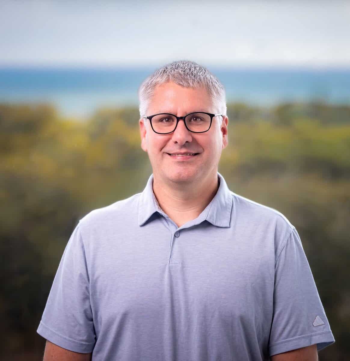 A man with short gray hair and glasses is wearing a light gray polo shirt, standing outdoors with blurred greenery and a body of water in the background.