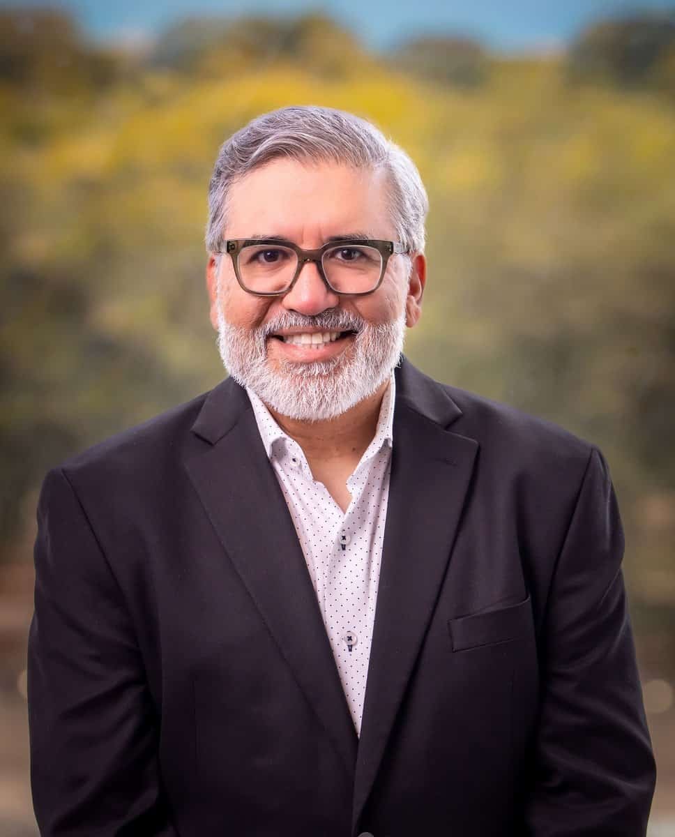 A man with gray hair, a beard, and glasses is wearing a black suit jacket over a white button-up shirt. He is smiling and standing in front of a blurred outdoor background with greenery.