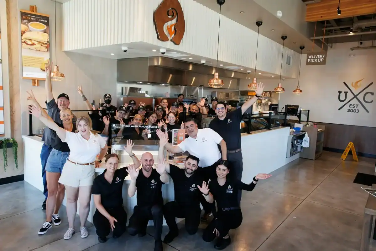 A large group of restaurant staff poses and smiles for a photo inside a modern eatery with copper light fixtures and a white counter, celebrating their achieved NPS and sales growth with BBI Survey Studio—Case Study: The Restaurant Group.