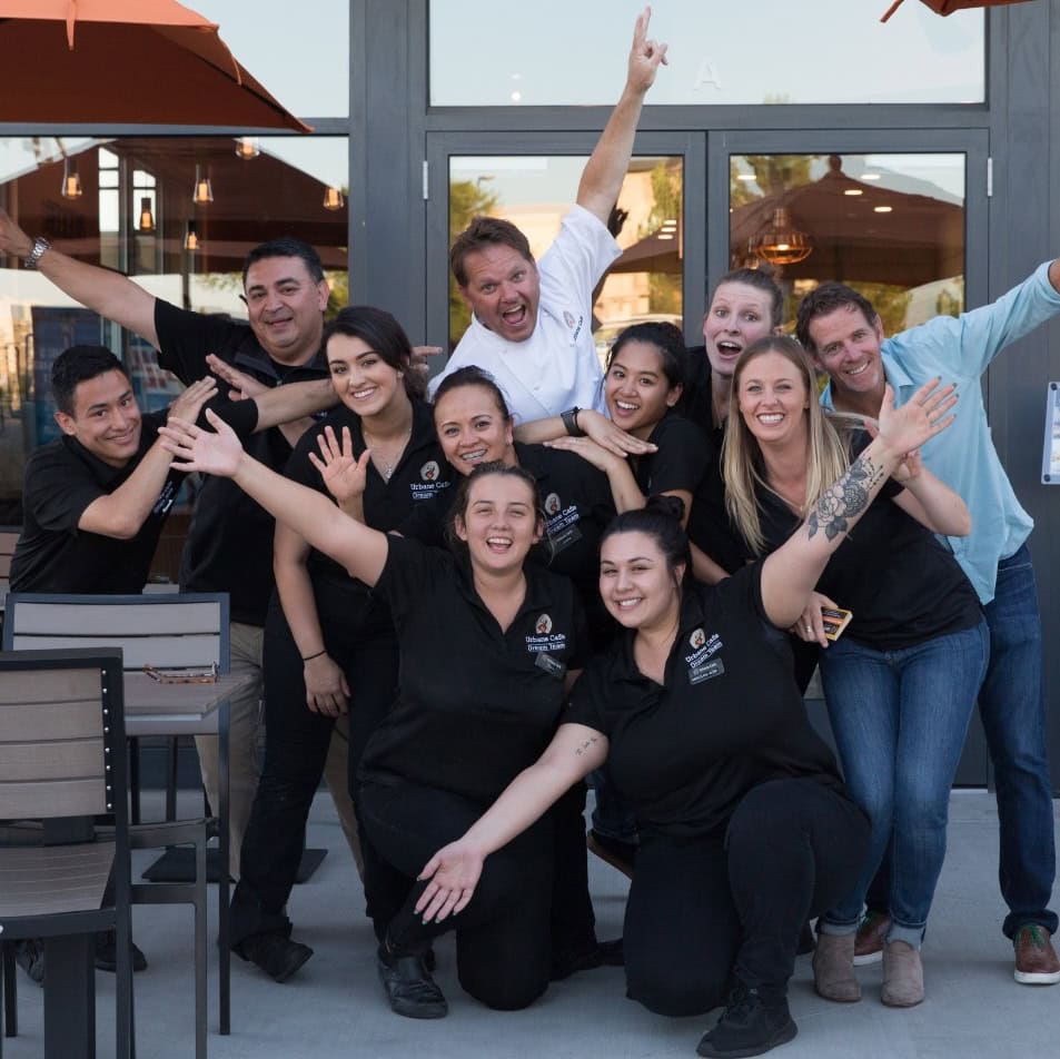A group of ten people pose energetically outside a building, most wearing matching black shirts. Celebrating their success as highlighted in Case Study: The Restaurant Group, several smile and gesture playfully at the camera.