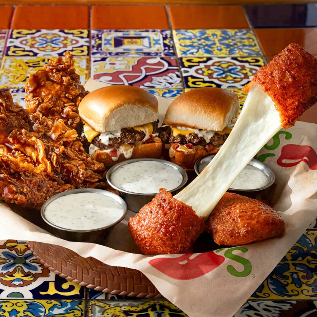 A basket contains crispy fried chicken, two cheeseburger sliders, mozzarella sticks with cheese stretching, and three cups of ranch sauce, set on a colorful tiled table—perfect for a Case Study:Urbane Cafe-inspired feast.