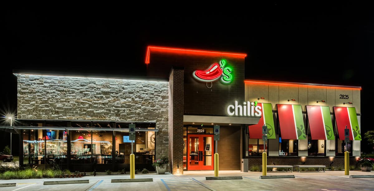 Exterior night view of a Chili’s restaurant with illuminated signage, red and green neon lights, striped awnings, and large windows showing the dining area inside—similar to Case Study: Urbane Cafe. Several parking spaces are visible in front.