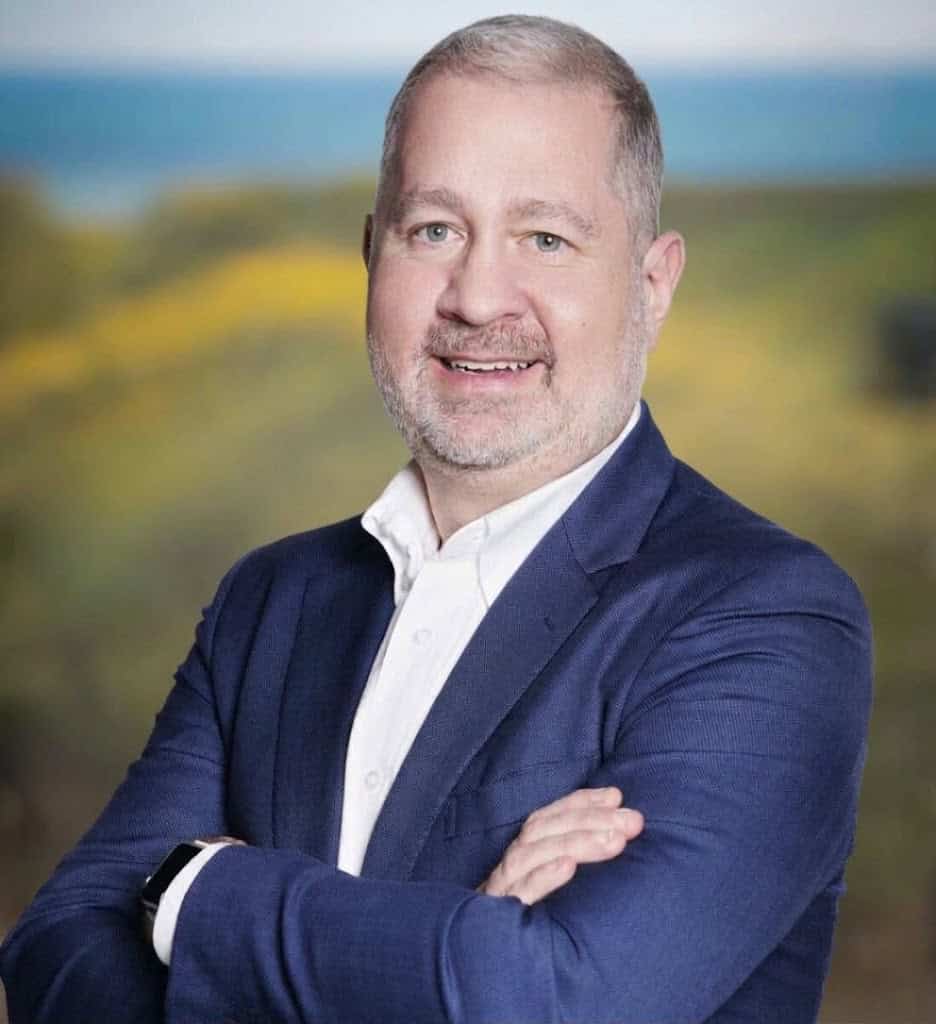 A middle-aged man with short hair and a goatee smiles while wearing a navy blazer and white dress shirt with his arms crossed.